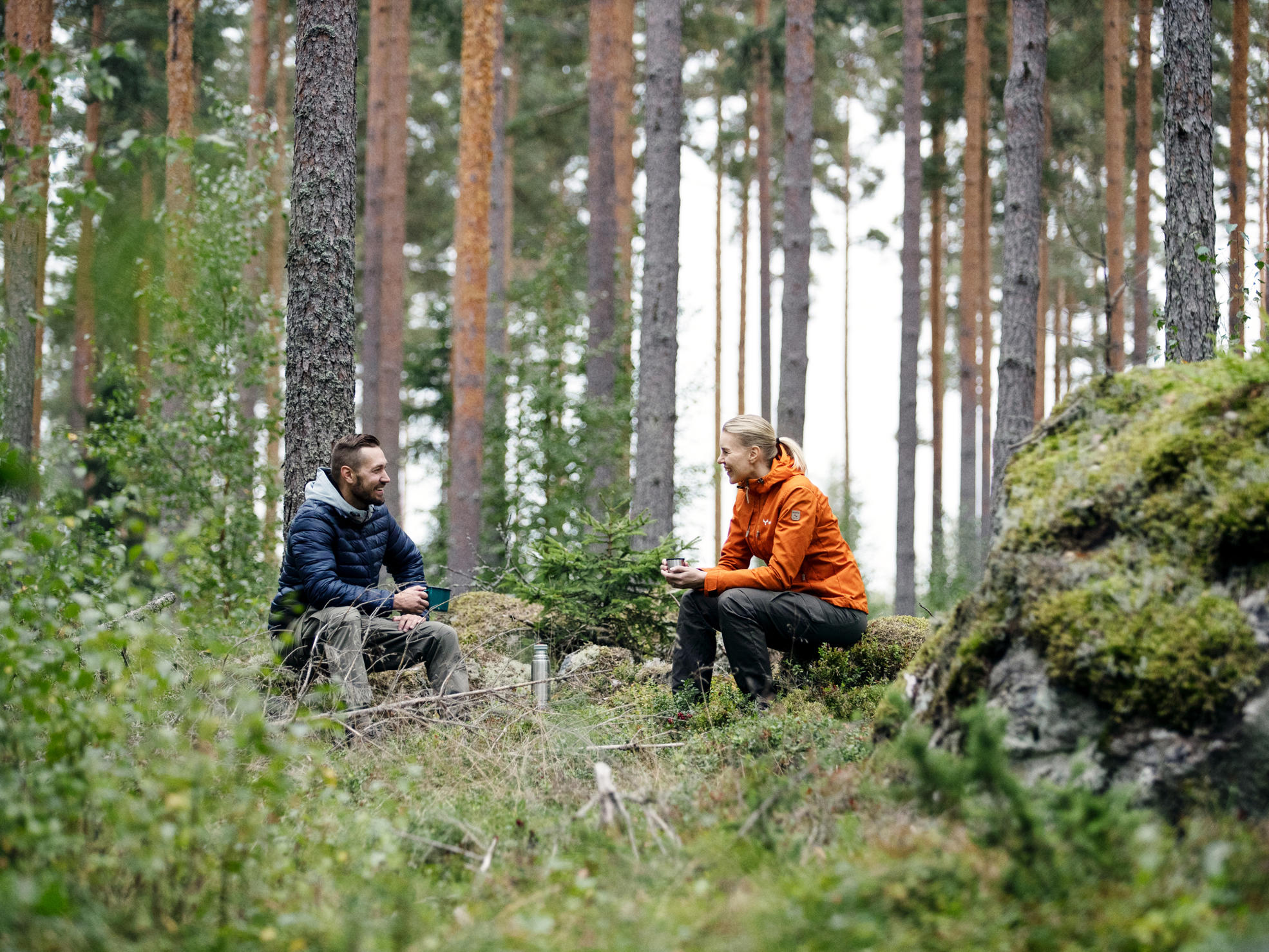 Metsänomistaja ja metsäasiantuntija istuvat metsässä juomassa kahvia ja juttelemassa.