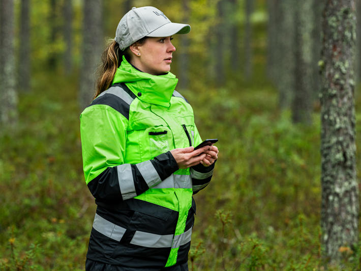 Metsa-asiantuntija seisoo metsässä puhelin kädessään ja arvioi ympäröivää metsää.
